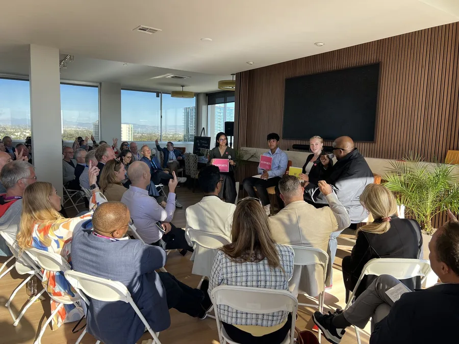 A panel of four facing an audience in a rooftop room with a city view. Panelists hold up pink Supports and yellow It Depends cards while audience members raise their hands.