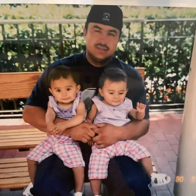 A baby photo of Marisol and another baby sitting on her dad's lap on a bench.