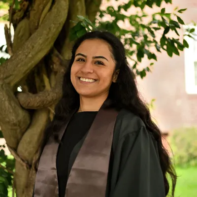 Marisol smiling in her graduation gown and Brown stole under a twisting tree.