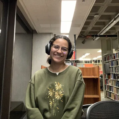 Marisol grinning in a library, wearing headphones and a green sweatshirt embroidered with flowers.